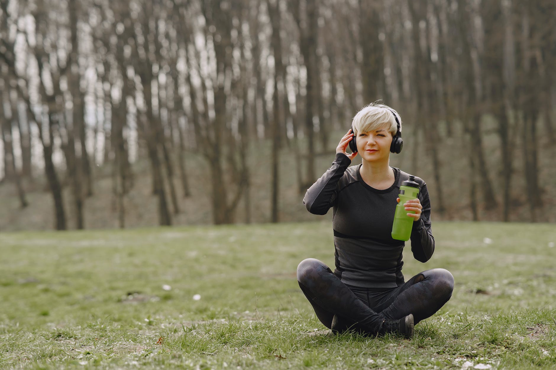young fit woman in headphones sitting on ground holding bottle of water and listening to music